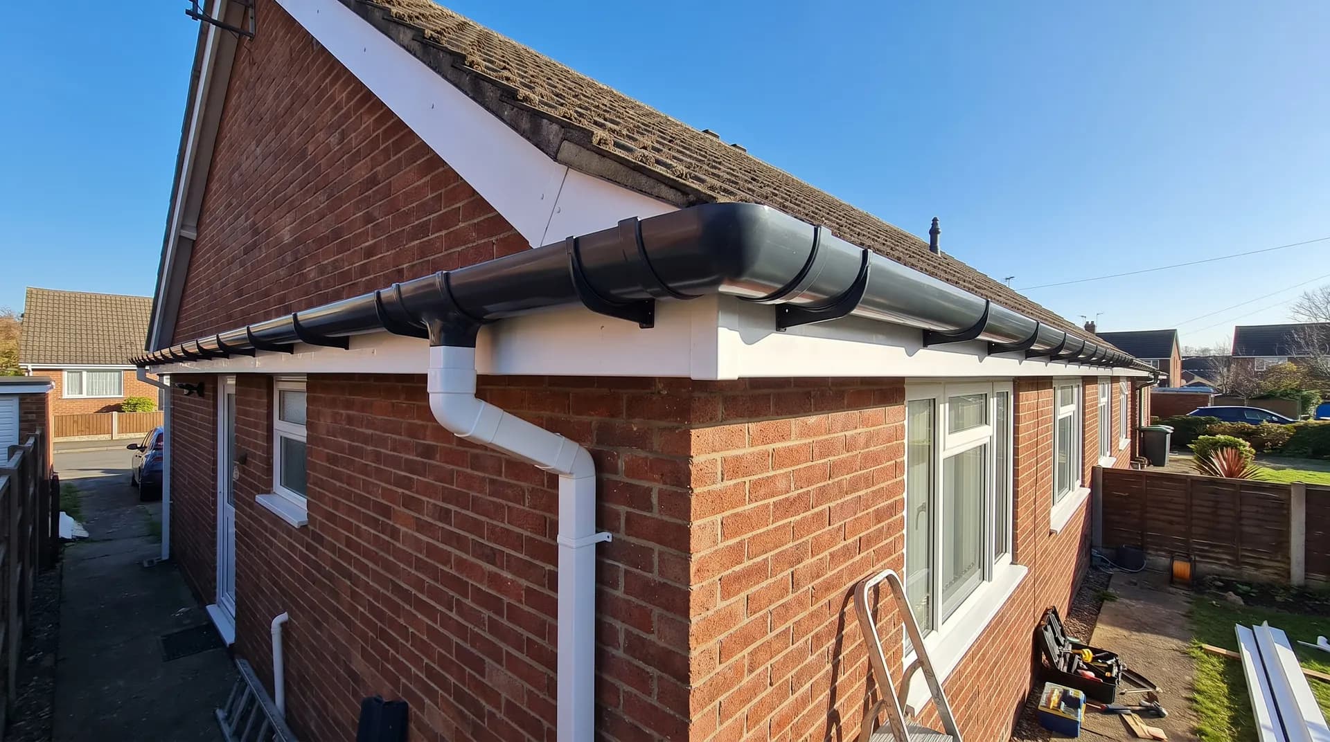 Dark grey half-round PVC guttering fitted along the eaves of a UK semi-detached house with white downpipe running down the wall to a hopper